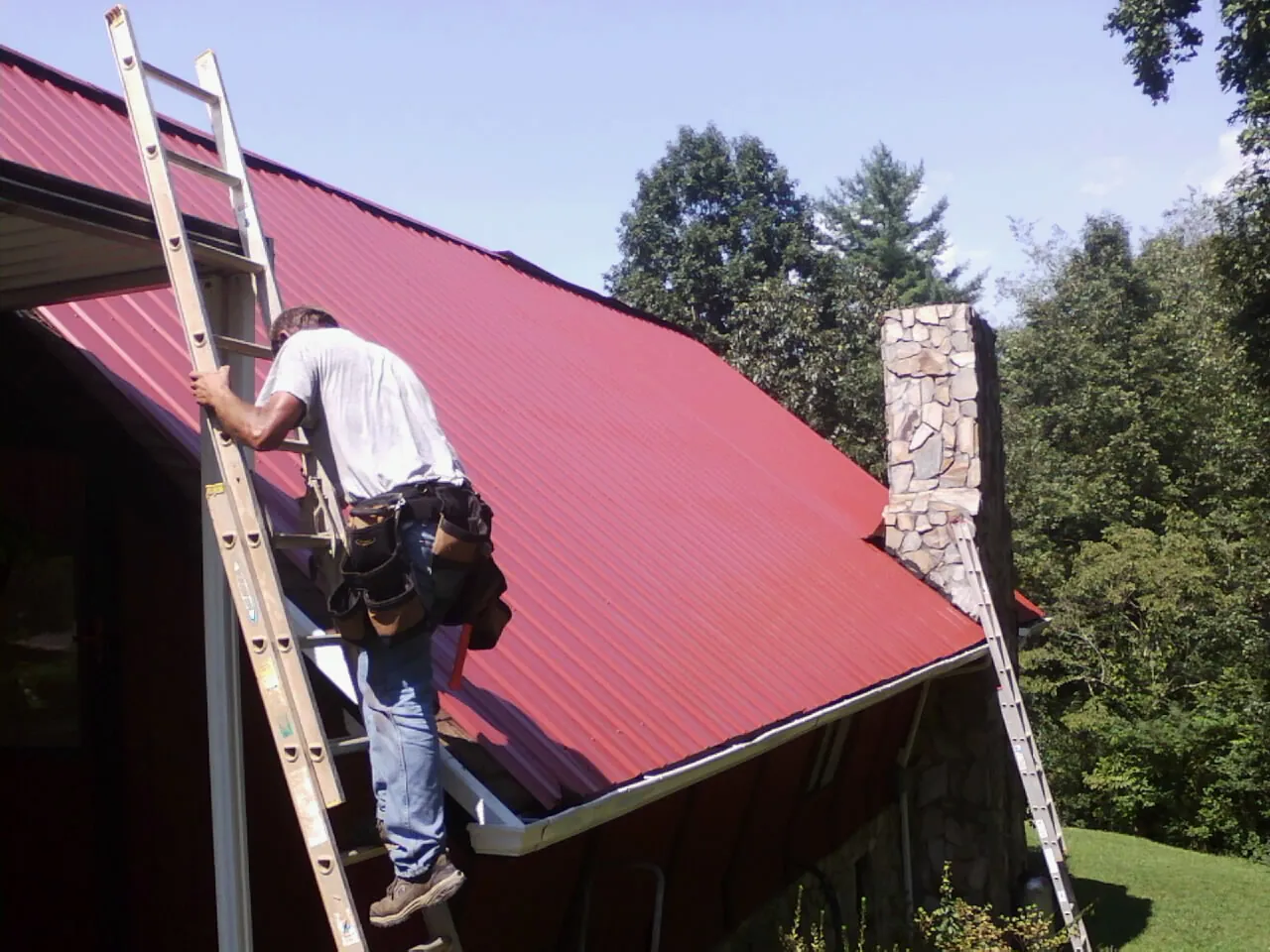 Roofer on a ladder beside a red metal roof and stone chimney — Soffit & Fascia Repair by Roofing Brothers in Sunbright